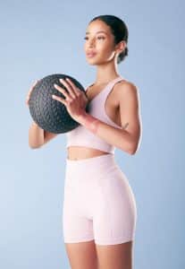 Young fitness woman standing with a black slam ball in studio against a blue background. 