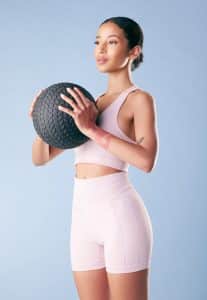 Young fitness woman standing with a black slam ball in studio against a blue background.