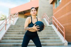 Sporty young woman working out using a black slam ball outdoors.