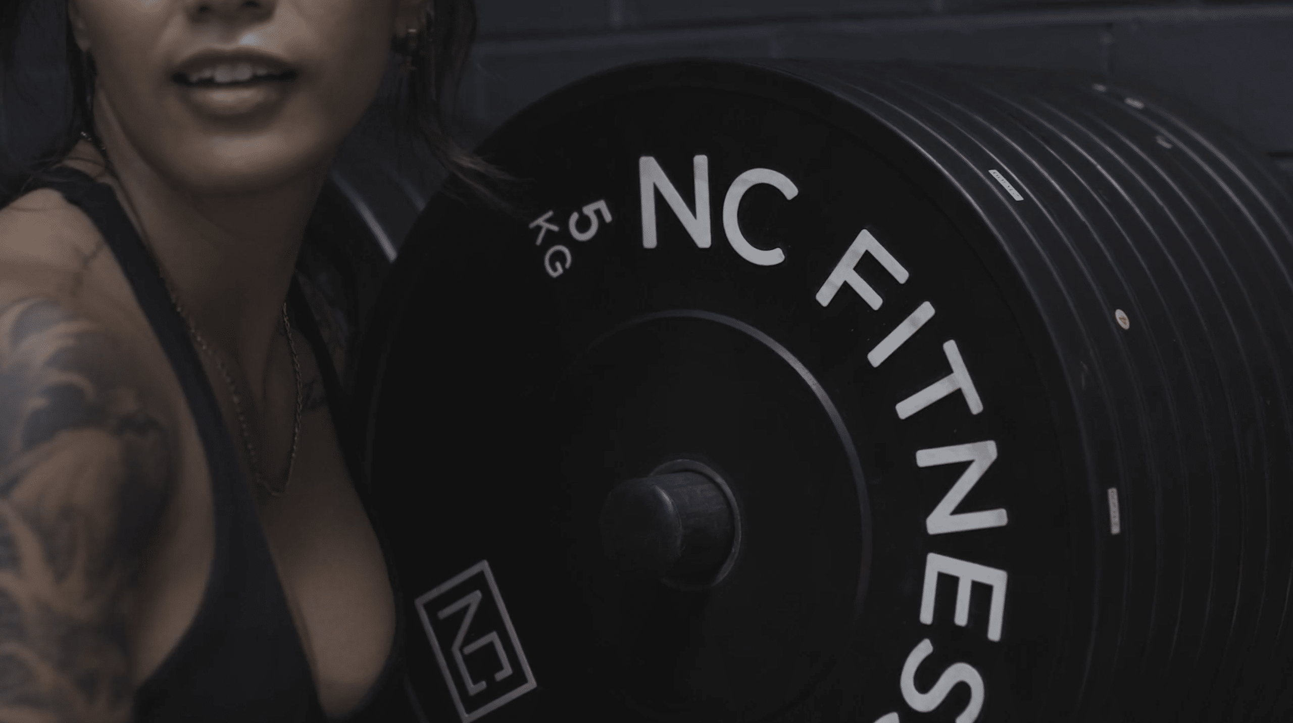 Tattooed woman in black sportswear next to a stack of 5kg black rubber NC Fitness Bumper Plates